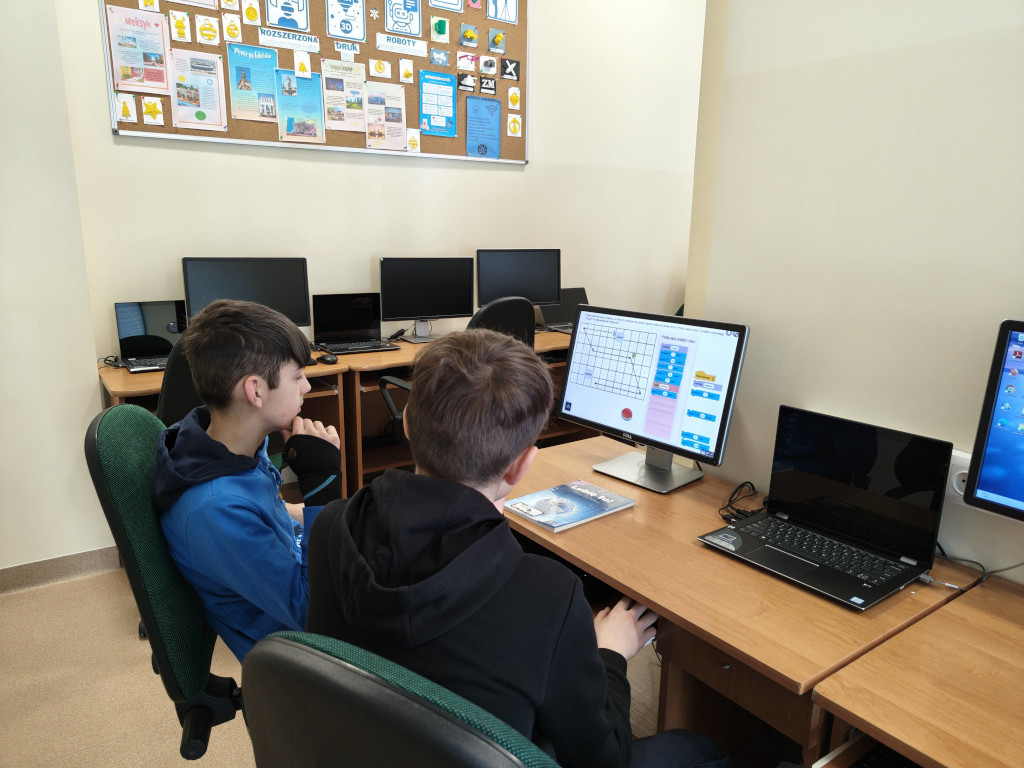 Uczniowie siedzą przy stanowiskach komputerowych w sali lekcyjnej i rozwiązują zadania w interaktywnych grach edukacyjnych dotyczących bezpieczeństwa w sieci.
