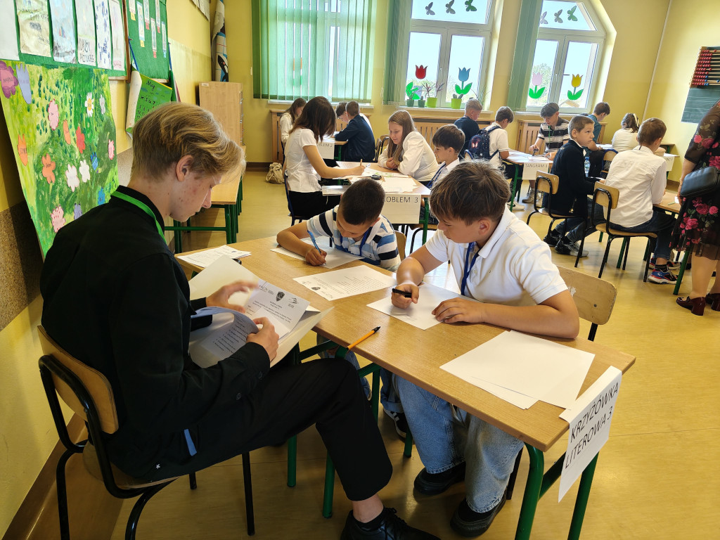Uczniowie siedzący przy szkolnych ławkach rozwiązują zadania matematyczne, a na pierwszym planie widoczny jest stolik z oznaczeniem „Krzyżówka literowa 3”.