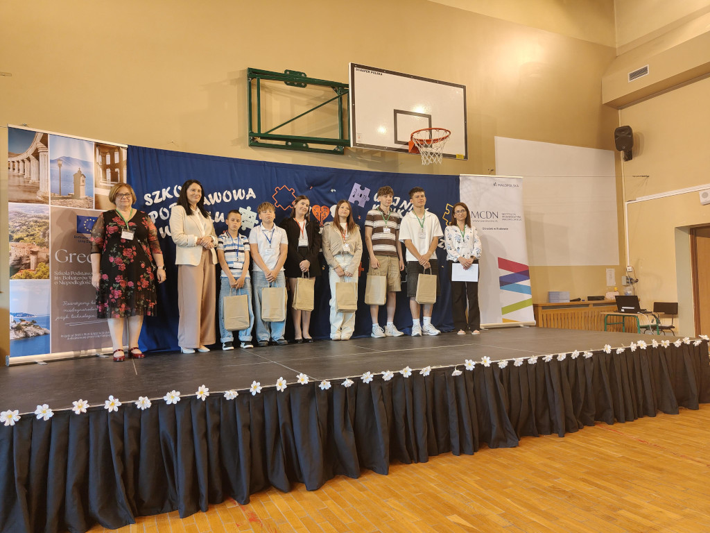 Uczestnicy konkursu oraz organizatorzy stoją na udekorowanej scenie w sali gimnastycznej podczas oficjalnego podsumowania wydarzenia.