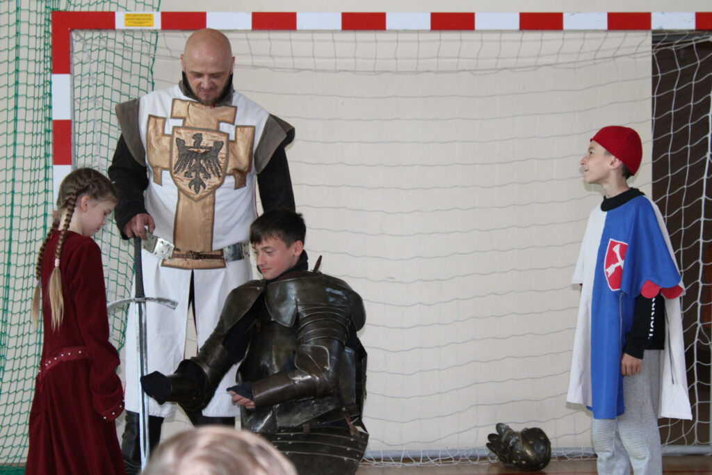 Do klęczącego w zbroi ucznia podchodzi uczennica w długiej bordowej sukni, podczas gdy rycerz i drugi uczeń obserwują sytuację.