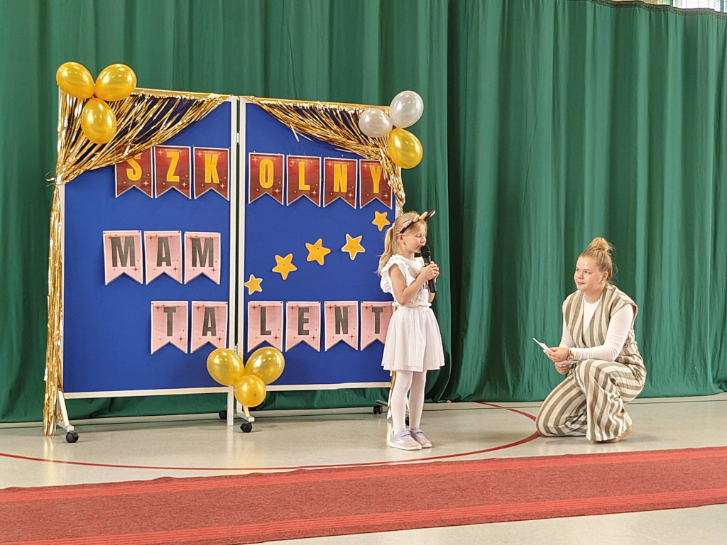 Mała dziewczynka w białej sukience i opasce z uszami śpiewa do mikrofonu podczas szkolnego konkursu talentów, a obok niej klęczy uśmiechnięta nauczycielka z kartką.