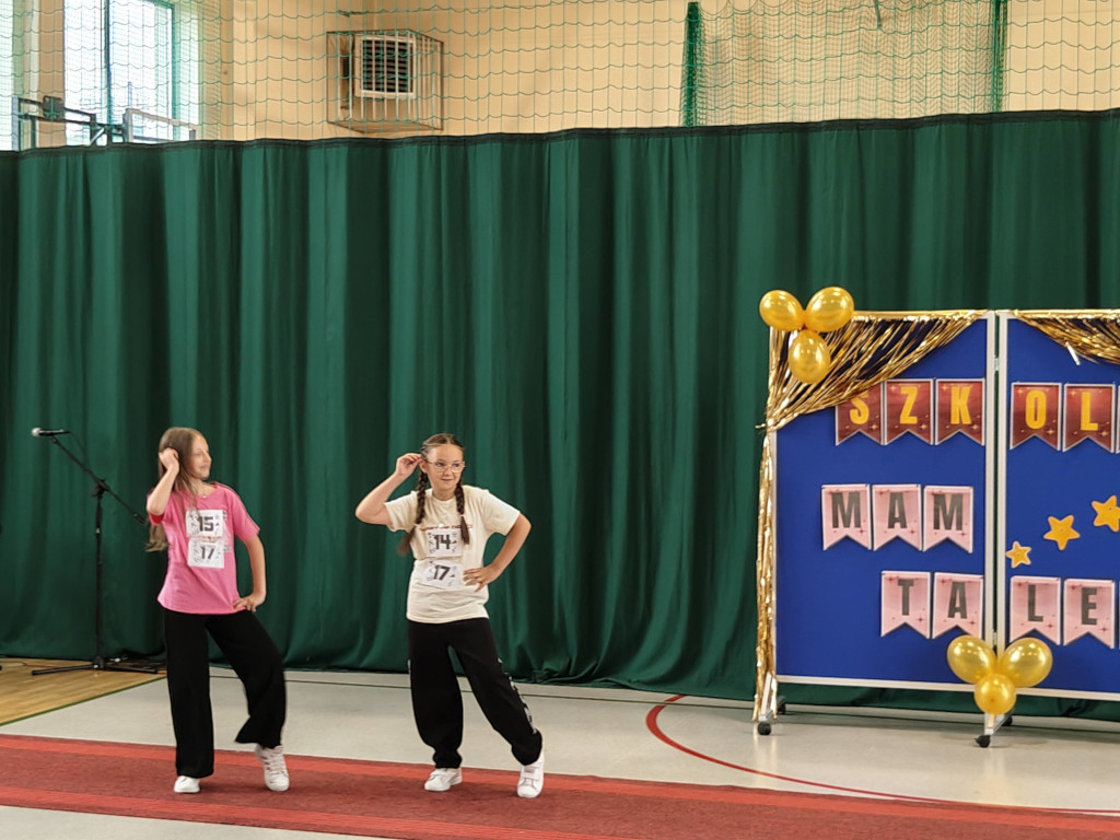 Dwie dziewczynki w luźnych strojach prezentują wspólny układ choreograficzny podczas szkolnego pokazu talentów.