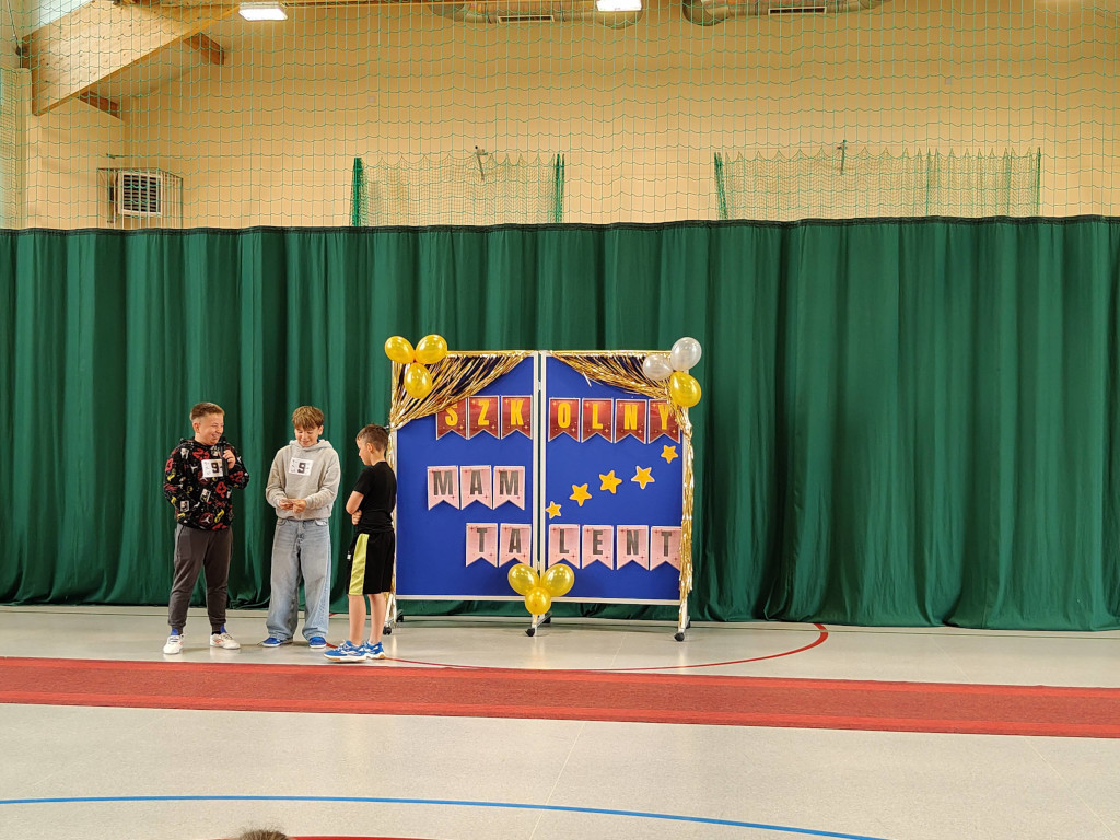Trzech chłopców w sportowych ubraniach stoi na czerwonym dywanie przed tablicą "Szkolny Mam Talent", a jeden z nich trzyma mikrofon.