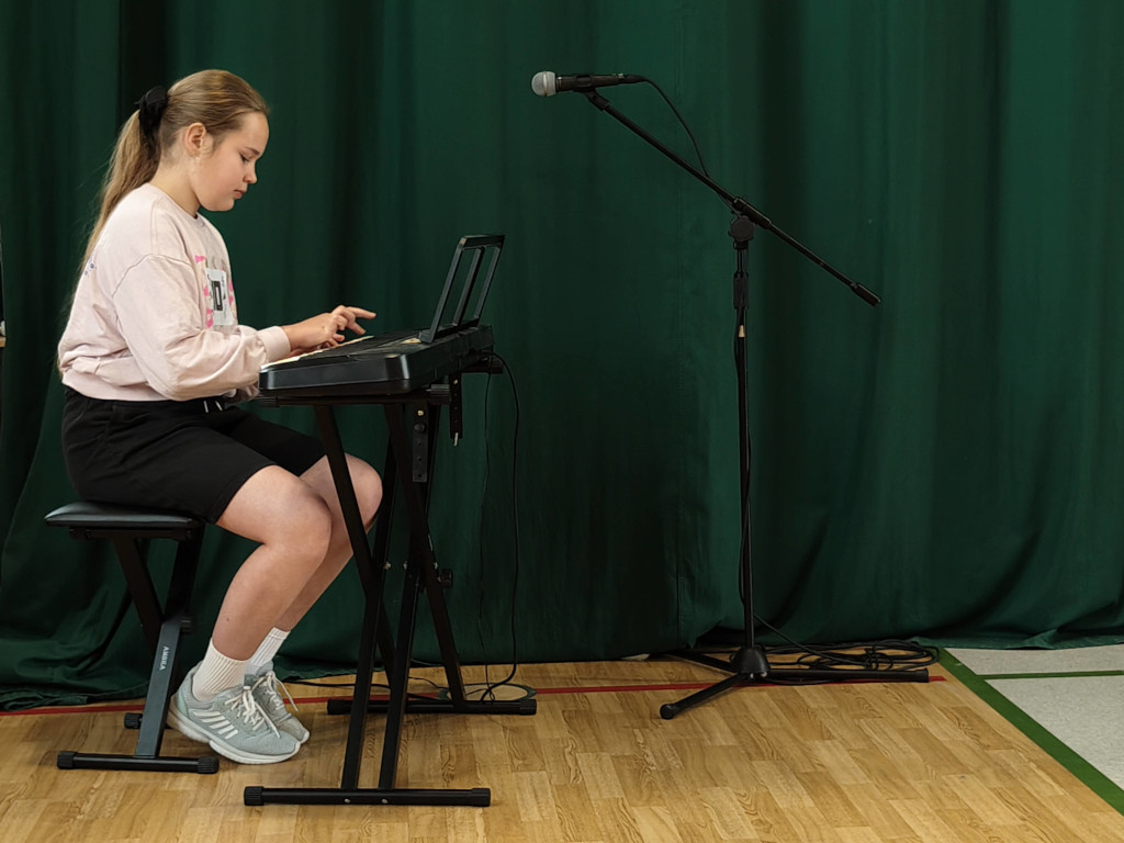 Uczennica siedzi na taborecie i gra na instrumencie klawiszowym ustawionym na statywie na tle długiej, zielonej kurtyny.