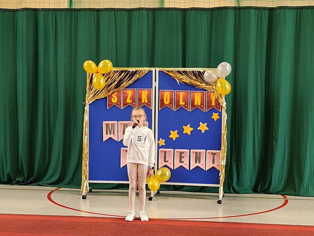 Dziewczynka w okularach i jasnej bluzie mówi do mikrofonu, stojąc na scenie przed niebieską tablicą z balonami.