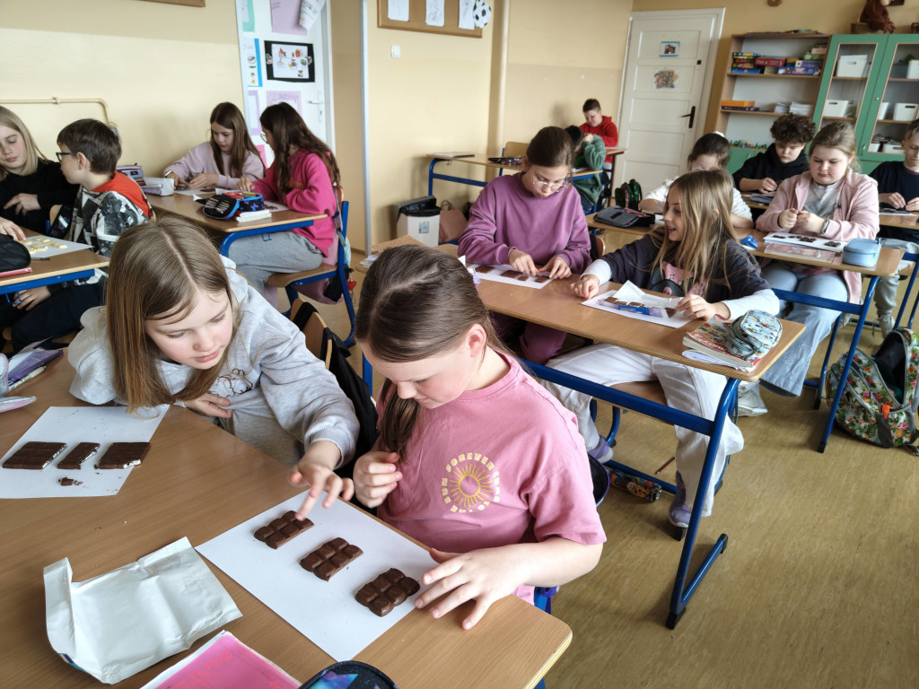 Klasa pełna uczniów pracujących w parach nad matematycznym zadaniem praktycznym z wykorzystaniem kostek czekolady.
