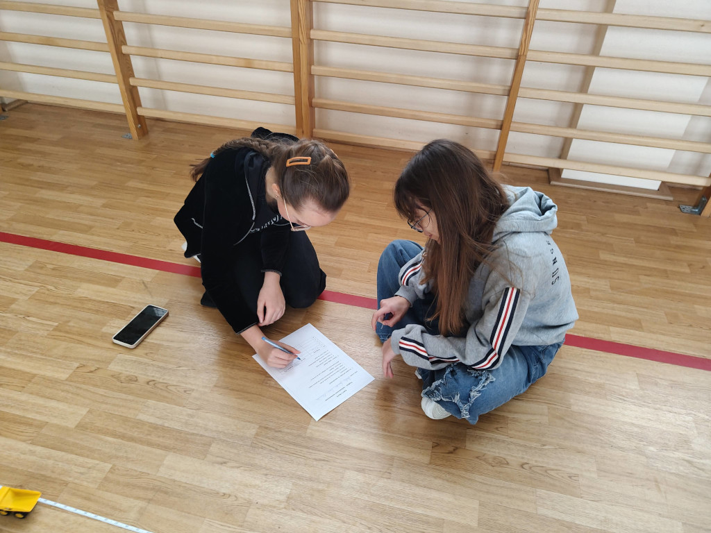 Dwie dziewczynki siedzą na podłodze sali gimnastycznej i wspólnie zapisują wyniki pomiarów na karcie pracy.