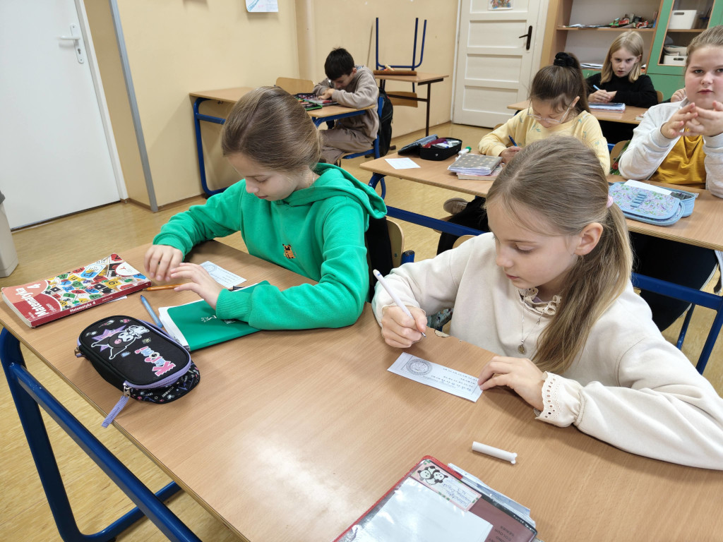 Dziewczynki w szkolnej ławce uważnie wypełniają podłużne, papierowe paski z zadaniami matematycznymi.