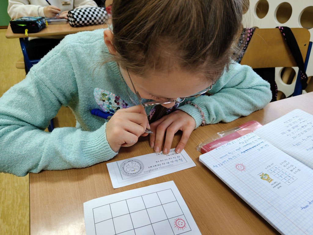 Dziewczynka w okularach i jasnoniebieskim swetrze w skupieniu rozwiązuje matematyczne zadanie na wąskim pasku papieru.