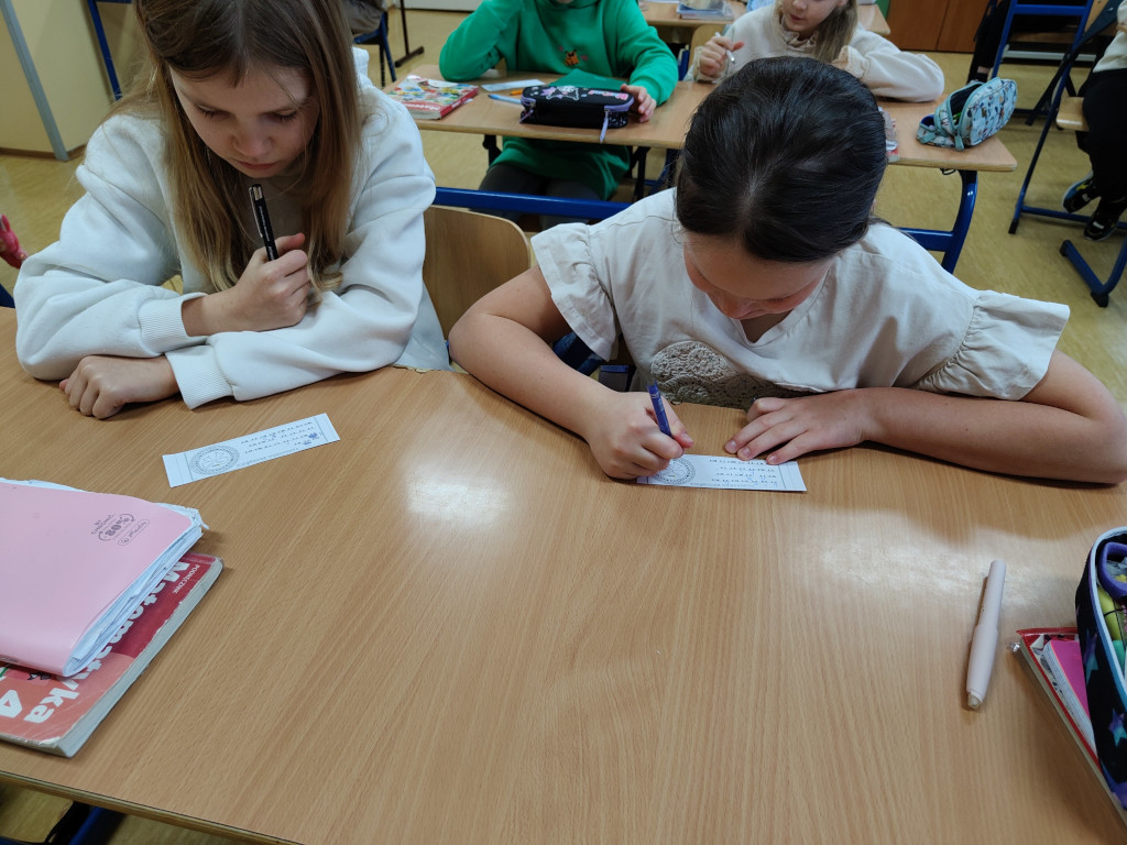 Dwie uczennice siedzące obok siebie w ławce piszą długopisami na papierowych paskach z ćwiczeniami matematycznymi.