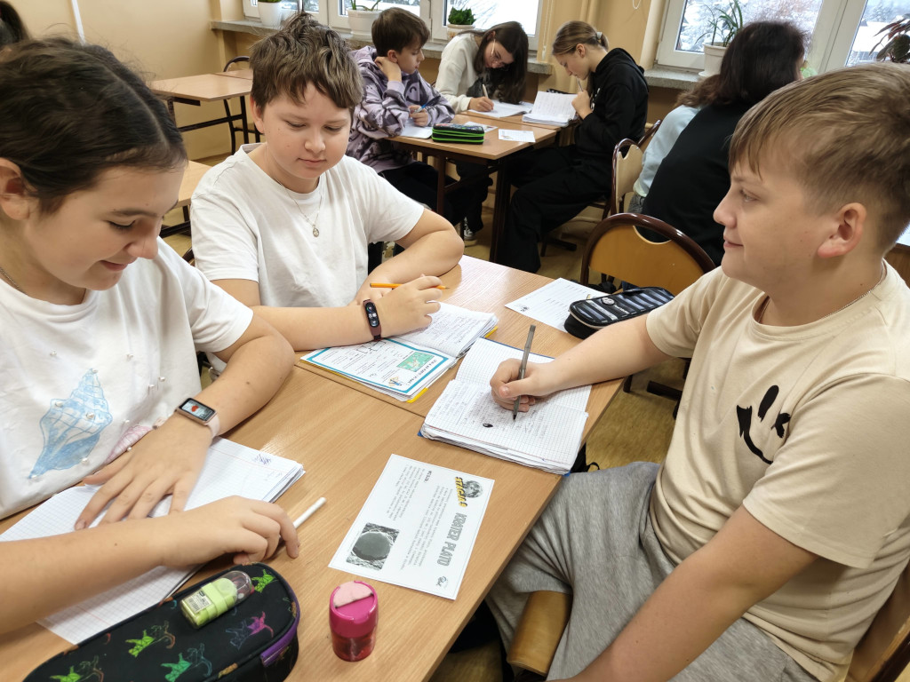 Trzej uczniowie siedzą przy szkolnej ławce i współpracują nad zadaniem, korzystając z otwartych zeszytów oraz karty pracy zatytułowanej „Krater Plato”.