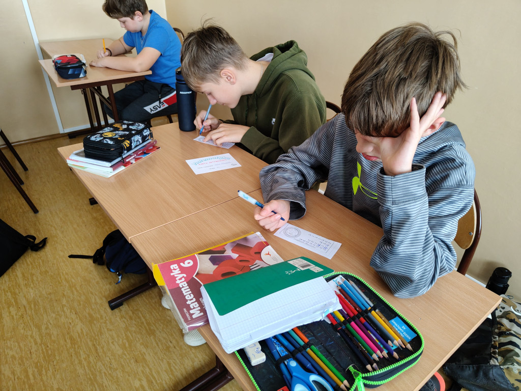 Trzej chłopcy w skupieniu rozwiązują zadania matematyczne w klasie lekcyjnej, a na pierwszym planie widać podręcznik do klasy 6.