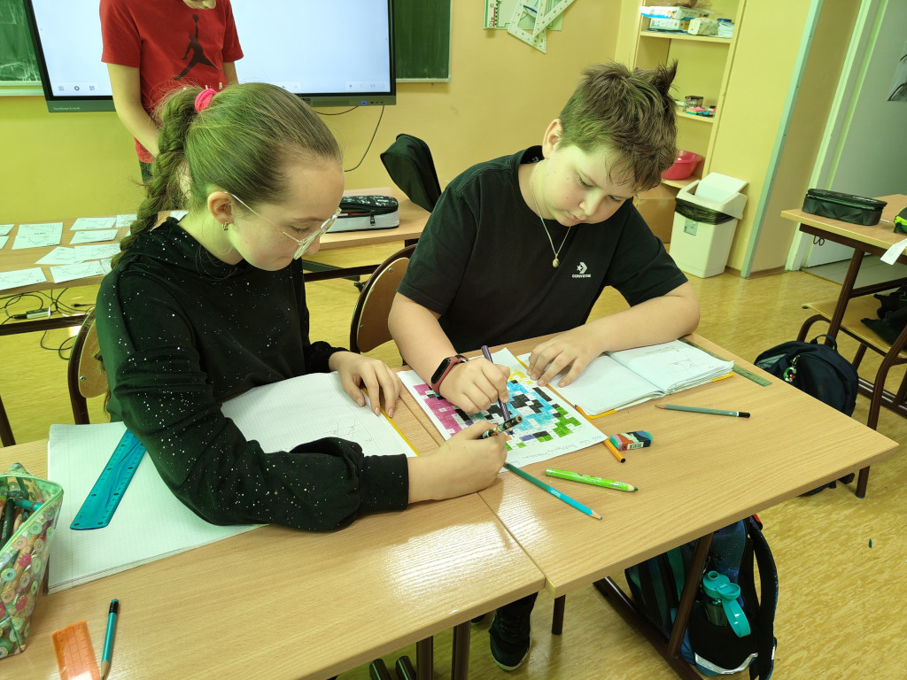 Uczennica i uczeń pracują wspólnie przy ławce, kolorując matematyczną mozaikę pikselową na karcie pracy.