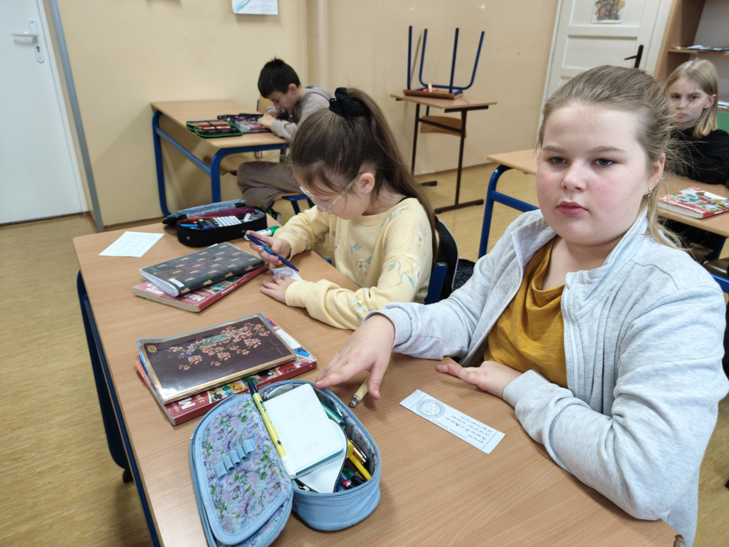 Dwie uczennice siedzą w szkolnej ławce, wypełniając długopisami wąskie paski papieru z zadaniami.