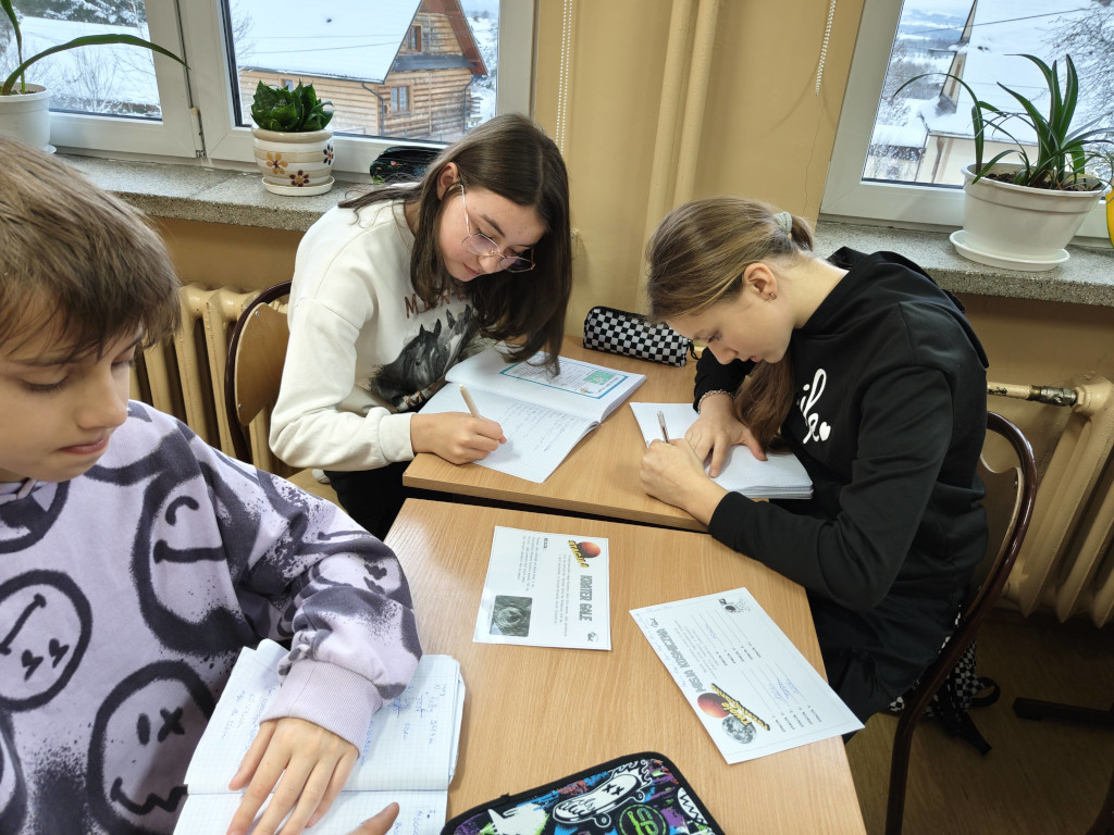 Dwie dziewczynki siedzą przy ławce pod oknem i uważnie piszą w zeszytach, rozwiązując zadania z karty „Misja Kosmiczna”.