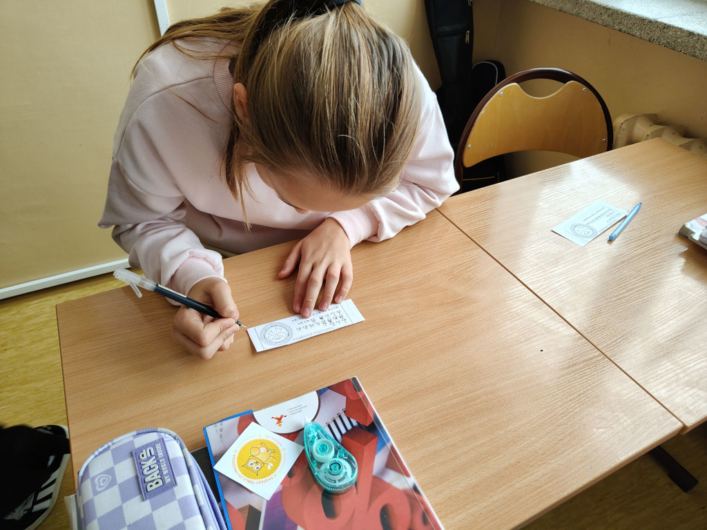 Dziewczynka pochyla się nad szkolną ławką, precyzyjnie wypisując odpowiedzi na podłużnym pasku papieru.