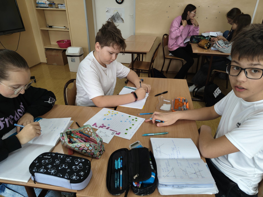 Grupa uczniów pracuje w ławkach nad zadaniami matematycznymi z wykorzystaniem rysunków geometrycznych i kolorowych kodów.