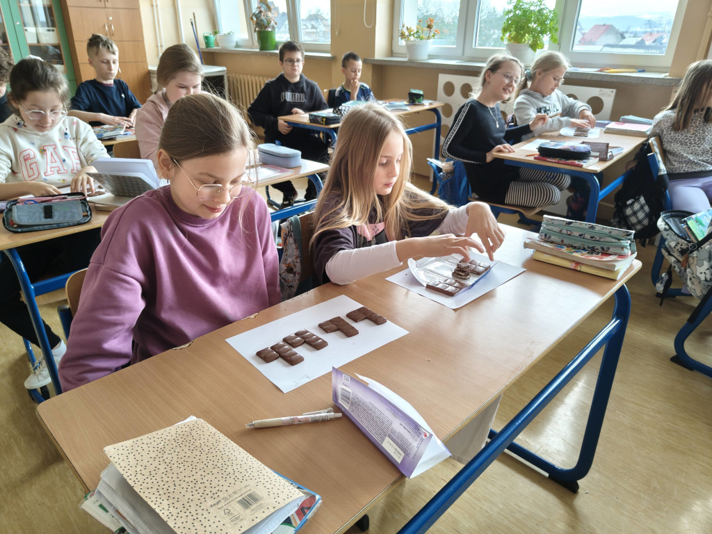 Uczennice siedzące w pierwszej ławce układają na białych kartkach części tabliczek czekolady podczas lekcji matematyki.