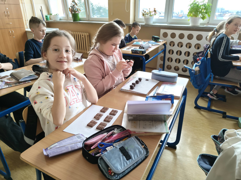 Uśmiechnięta dziewczynka w okularach pozuje przy szkolnej ławce, na której ułożone są kostki czekolady służące do nauki ułamków.