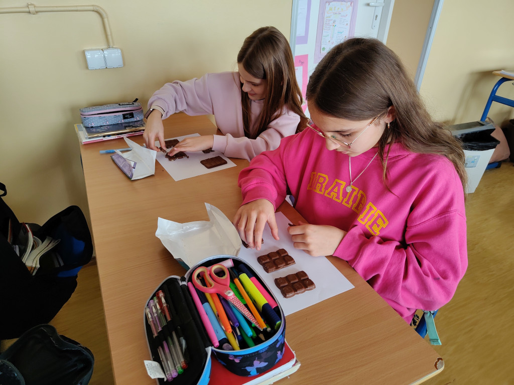 Dwie dziewczynki pracują wspólnie przy szkolnej ławce, układając kostki mlecznej czekolady na czystej kartce.