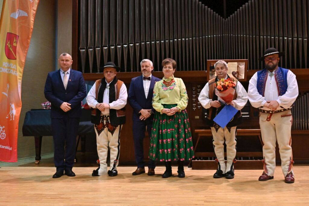 Pamiątkowe zdjęcie sześciu osób na scenie. Widoczni są mężczyźni w garniturach oraz przedstawiciele regionu w strojach ludowych (górale i góralka) na tle piszczałek organowych.