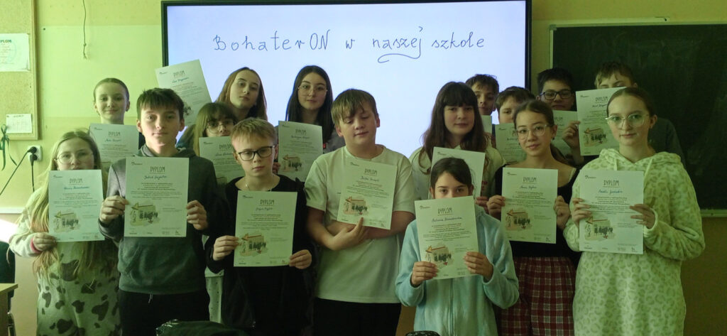 Grupa uśmiechniętych uczniów stoi w klasie i prezentuje otrzymane dyplomy na tle interaktywnej tablicy z napisem „BohaterON w naszej szkole”.