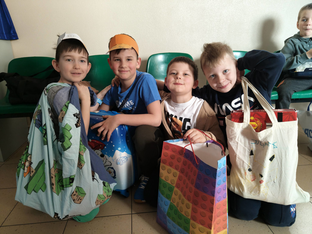 Czterej chłopcy kucają blisko siebie, prezentując swoje kreatywne zamienniki plecaków: niebieską walizkę kabinową, kolorową papierową torbę w klocki, płócienną torbę z rysunkiem oraz poszewkę na poduszkę.