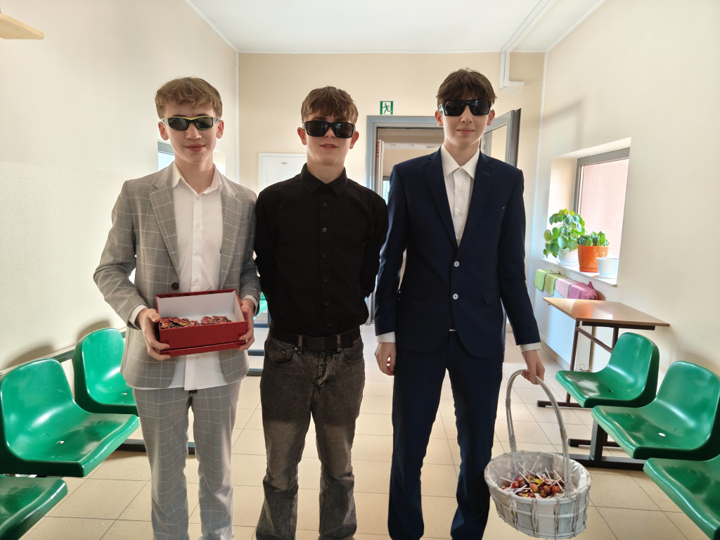 Trzej elegancko ubrani uczniowie w okularach przeciwsłonecznych stoją na korytarzu, trzymając pudełko i koszyk z drobnymi upominkami.