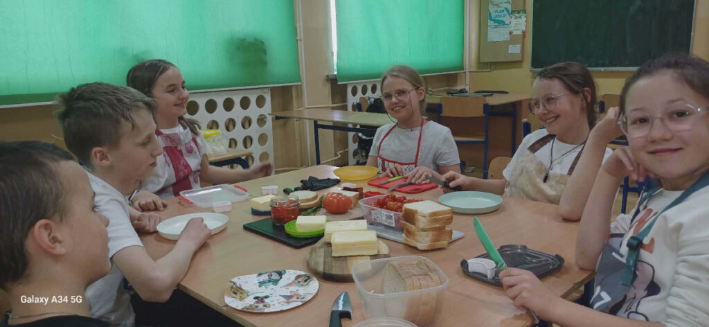 Uśmiechnięci uczniowie siedzą wspólnie przy stole pełnym składników na kanapki w trakcie szkolnych zajęć kulinarnych.