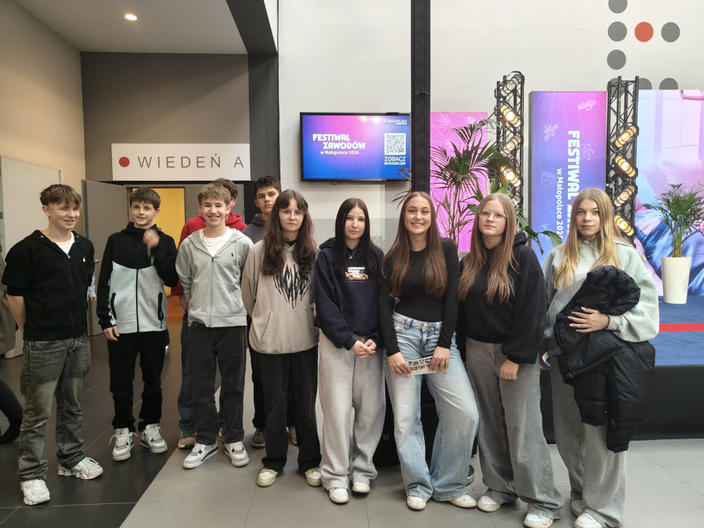 Grupa młodych osób stoi obok siebie, pozując do pamiątkowego zdjęcia na tle ekranów z wyświetlonym logo „Festiwal Zawodów w Małopolsce 2026”.