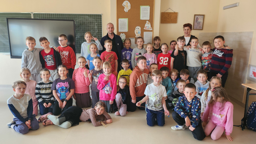 Uczniowie pozują do pamiątkowej fotografii przy ławkach szkolnych w otoczeniu nauczycieli i zaproszonego gościa