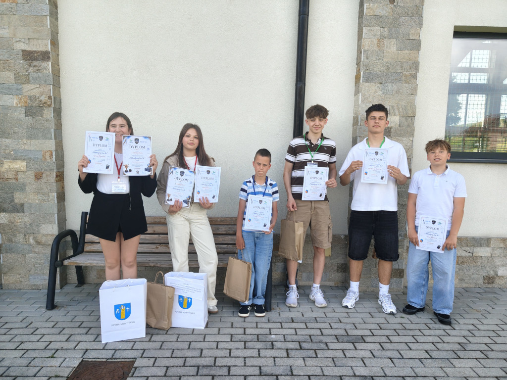 Sześcioro uczniów stoi przed budynkiem szkoły, prezentując zdobyte dyplomy oraz torby z upominkami.