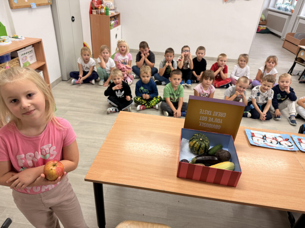 Przedszkolaki oczekują na lekcję o zmysłach. Zdjęcie przedstawia pudełko pełne owoców i warzyw.