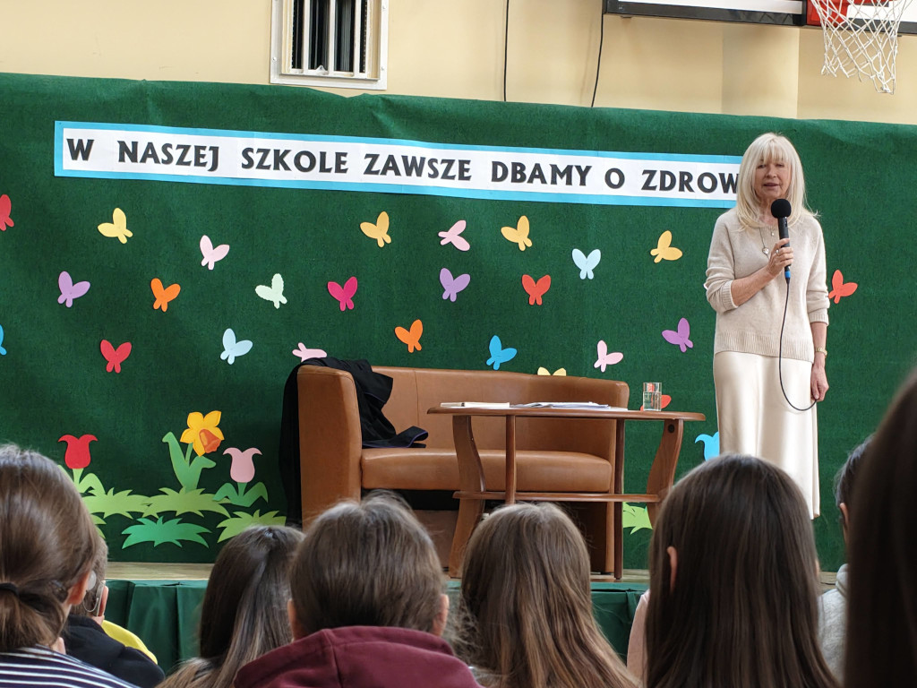 Blondwłosa prelegentka mówi do mikrofonu ze sceny w sali gimnastycznej, patrząc na zgromadzonych w pierwszym planie słuchaczy.